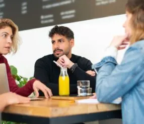 Quatro pessoas sentadas em uma mesa conversando em um café, com bebidas sobre a mesa.