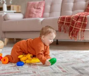 Brinquedos para bebê de 8 meses: o que observar antes de comprar Bebê em trajes laranja brincando no chão com brinquedos coloridos.